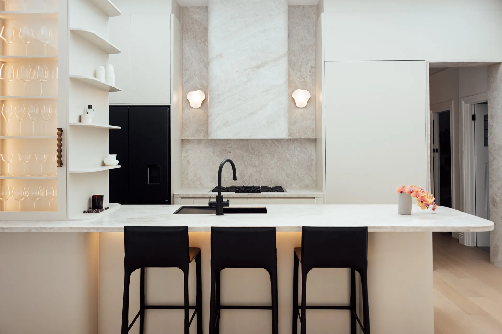 Wide kitchen island with three stools, illuminated glass shelving and butler pantry beyond.